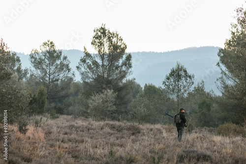 Hunter holding a gun while standing outdoors in the mountain forest looking for a prey.