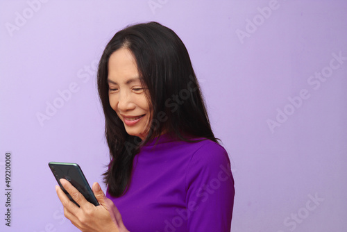 Portrait of an Asian model holding a mobile phone Smiling, wearing a purple shirt against a light purple background.