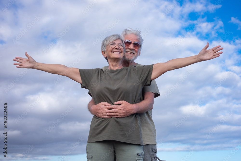Happy senior man hugging his wife while she stands with open arms. Beautiful smiling white-haired elderly couple enjoying retirement and freedom on outdoor excursion Happy senior man hugging his wife while she stands with open arms. Beautiful smiling white-haired elderly couple enjoying retirement and freedom on outdoor excursion