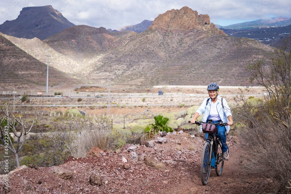 Cheerful senior woman riding in a country road with bike, cyclist wearing sport helmet. Mountains and cloudy sky in background Cheerful senior woman riding in a country road with bike, cyclist wearing sport helmet. Mountains and cloudy sky in background
