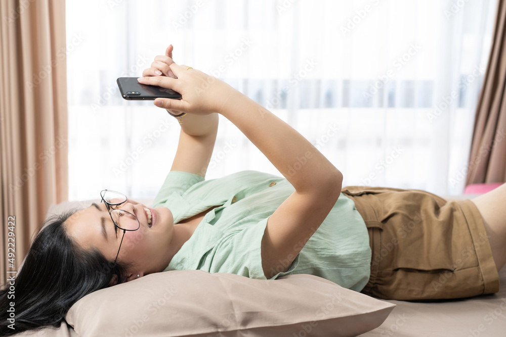 Beautiful glasses Asian woman lie on her back, chatting, typing, and texting, mobile in her bedroom with a pastel green - brown color theme. Beautiful glasses Asian woman lie on her back, chatting, typing, and texting, mobile in her bedroom with a pastel green - brown color theme.