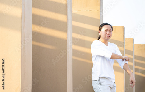 Asian long hair, Man in white long arm t-shirt is standing at the rooftop outdoor field with light brown wall bacgkround.