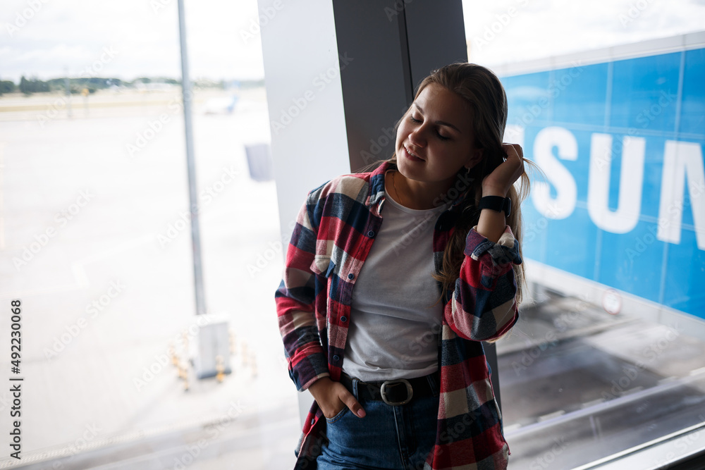 Obraz premium A beautiful attractive ukrainian woman with blond hair is standing by a large window, wearing momom jeans and a t-shirt with a plaid shirt. Girl in casual comfortable clothes