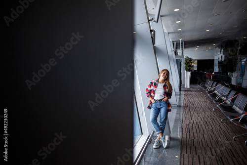 A beautiful attractive ukrainian woman with blond hair is standing by a large window  wearing momom jeans and a t-shirt with a plaid shirt. Girl in casual comfortable clothes
