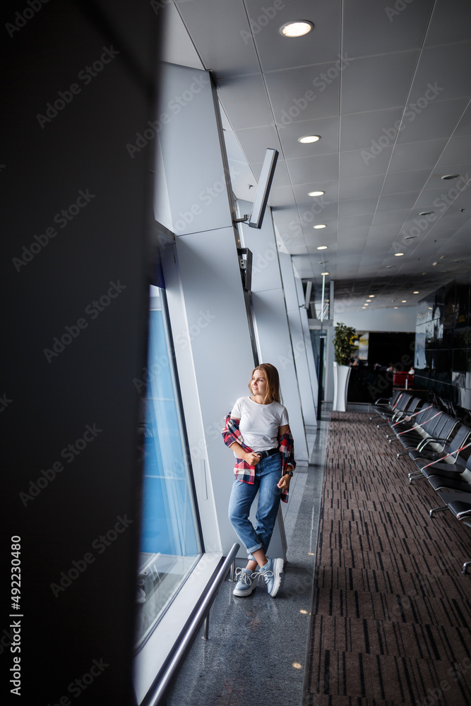 A beautiful attractive ukrainian woman with blond hair is standing by a large window, wearing momom jeans and a t-shirt with a plaid shirt. Girl in casual comfortable clothes