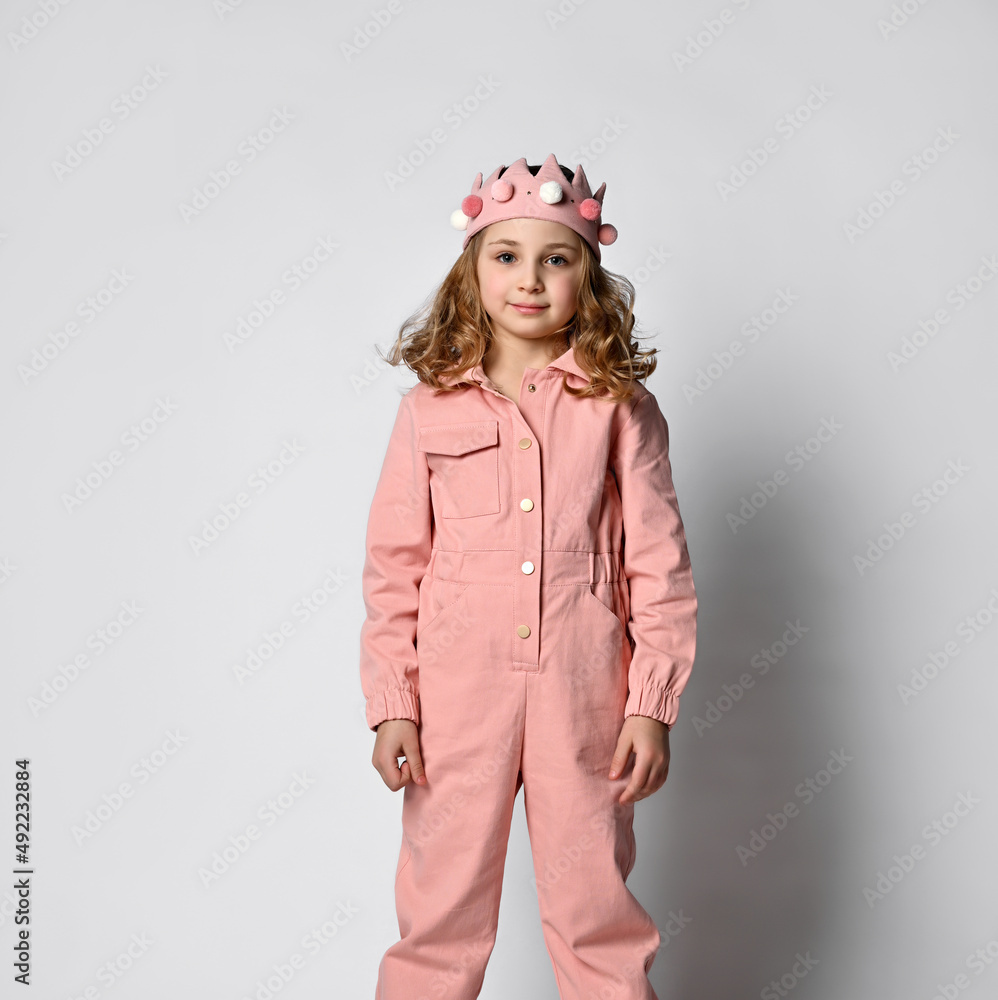 cute girl with a plush crown, standing upright in a dusty rose cotton summer jumpsuit with comfortable pockets. on a light background. cute girl with a plush crown, standing upright in a dusty rose cotton summer jumpsuit with comfortable pockets. on a light background.