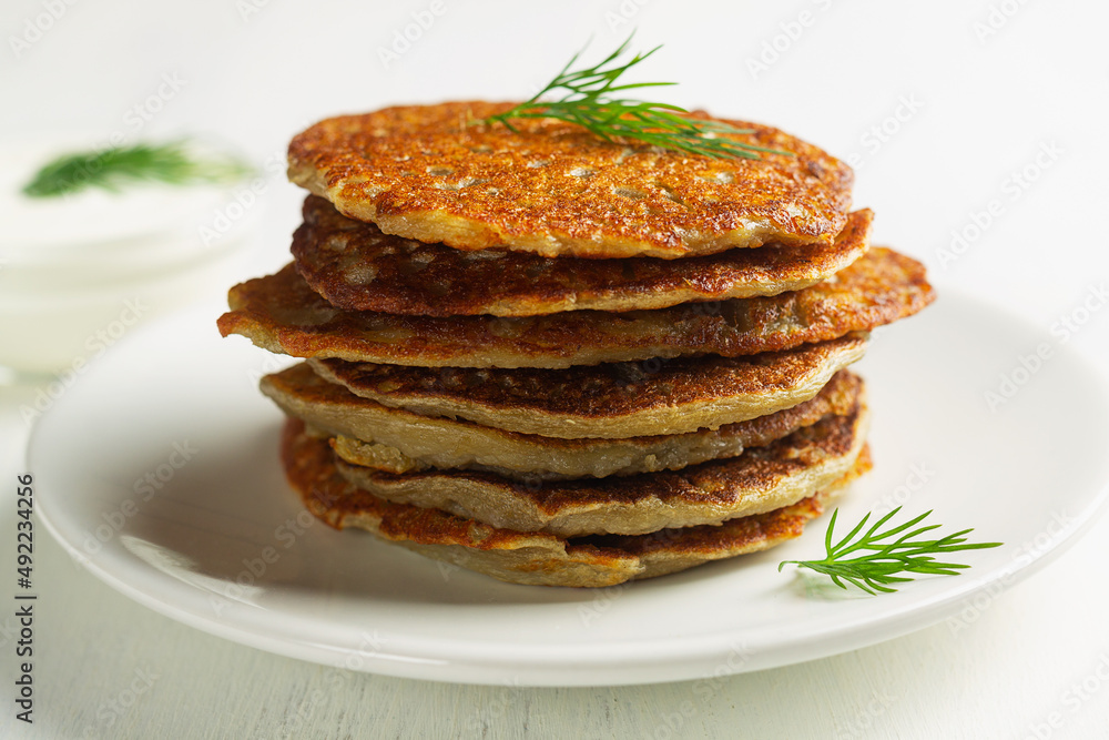stacks of toasted Potato pancakes with white sour cream, green herbs on the white plate. Belarusian cuisine. close-up potato pancakes on the white background with copy space. stacks of toasted Potato pancakes with white sour cream, green herbs on the white plate. Belarusian cuisine. close-up potato pancakes on the white background with copy space.