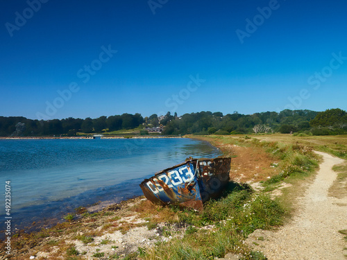 A derelict, abandoned and corroded boat sits beside the waters of Bembridge Harbour under a bright blue, summery sky.