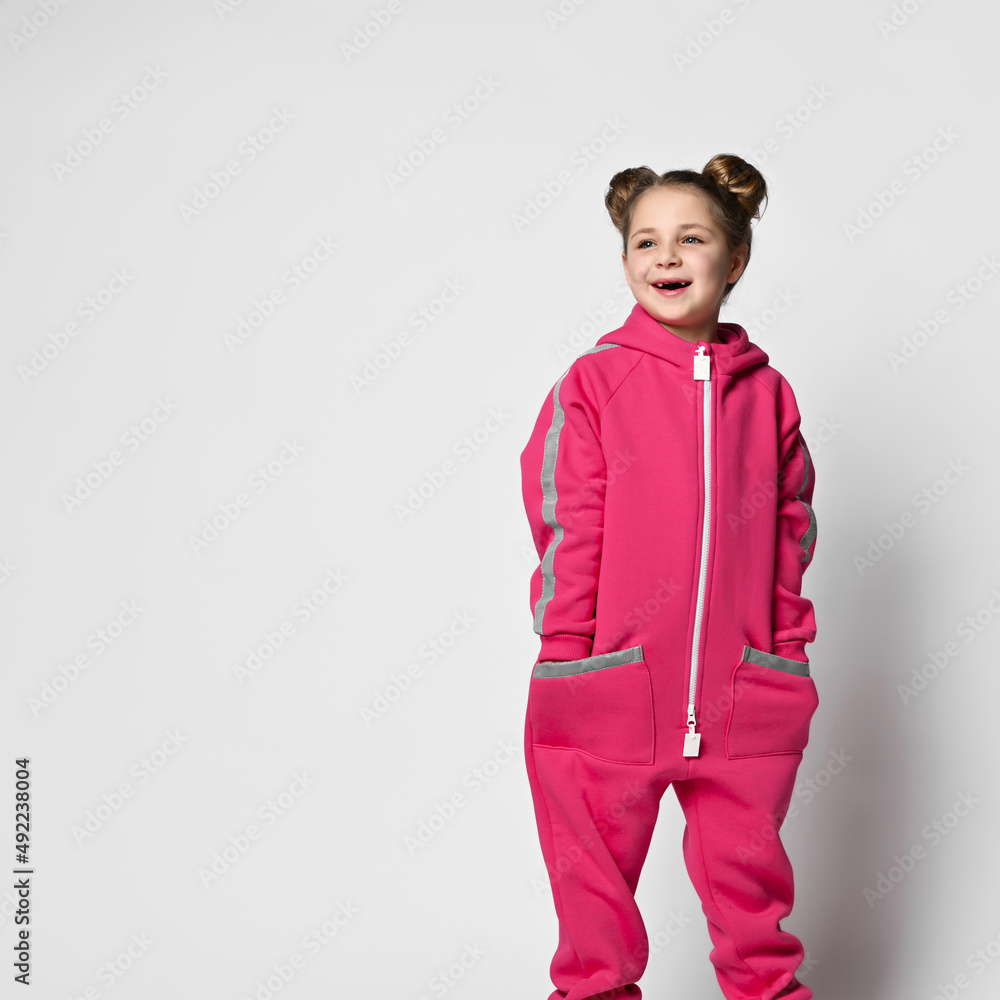 smiling girl in a pink modern jumpsuit with reflective safety stripes on the sleeves and pockets stands with her hands in her pockets and looks to the side against a light background. smiling girl in a pink modern jumpsuit with reflective safety stripes on the sleeves and pockets stands with her hands in her pockets and looks to the side against a light background.
