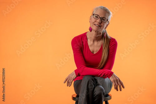 Beautiful smiling happy blond mature woman wearing eyeglasses on solid orange background with copy space sitting on stool. © Robert Peak