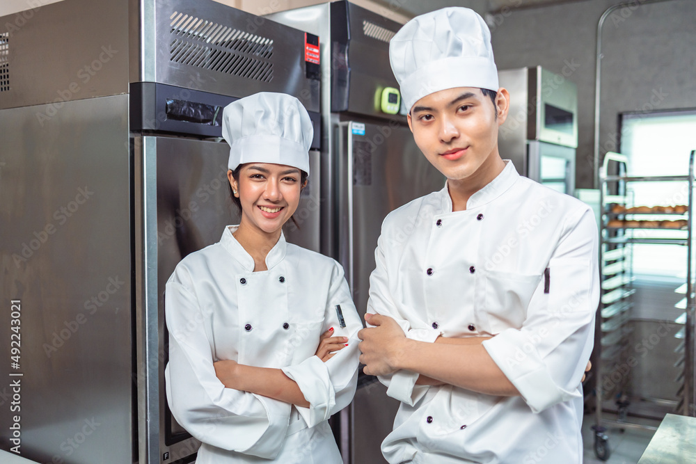 Couple asian young female bakers looking at camera..Chefs  baker in a chef dress and hat, cooking together in kitchen.Team of professional cooks in uniform preparing meals for a restaurant.