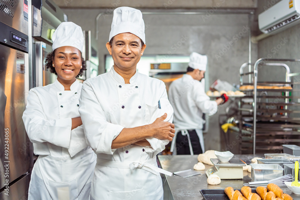 Young  African  female and  senior asian male bakers looking at camera..Chefs  baker in a chef dress and hat, cooking together in kitchen.Team of professional cooks in uniform.