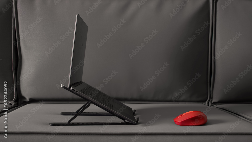 Steel thin laptop on a stand and a red computer mouse on the surface of a  gray corner loft sofa. The concept of remote work and Internet access from  home. Nobody Stock