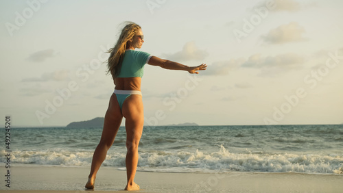 Sexy woman walking alone on beach coastline and relax warm golden sunset. Woman walking on water in blue swimsuit and sunglasses. Concept rest tropical resort traveling tourism happy summer holidays