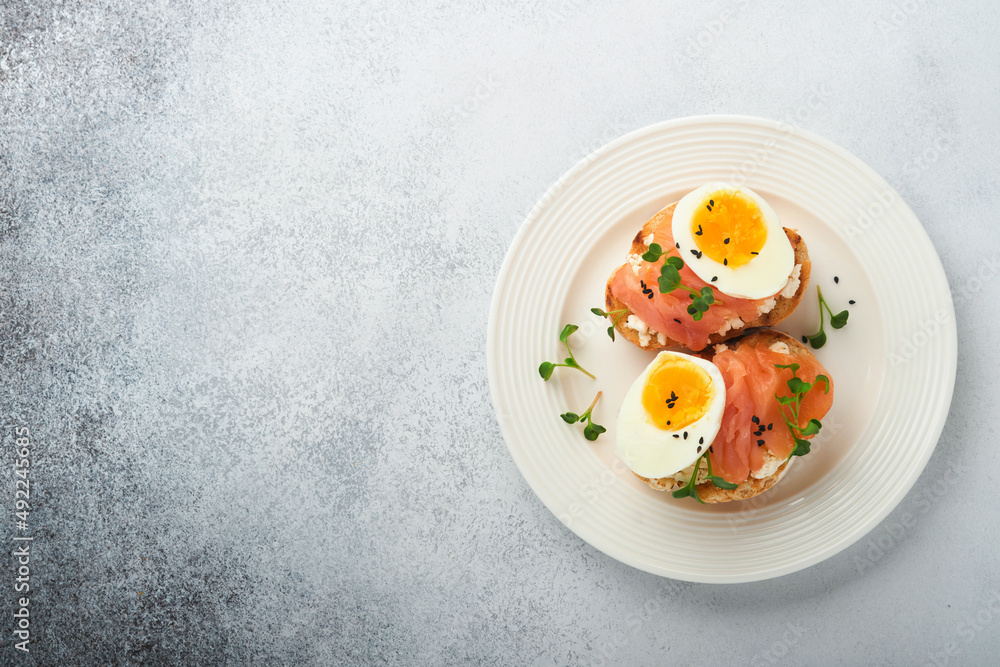 Sandwich with delicious toppings on top: smoked salmon, eggs, herbs and microgreens radish, black sesame seeds over white plate on concrete background. Healthy open sandwich superfood. Top view. Sandwich with delicious toppings on top: smoked salmon, eggs, herbs and microgreens radish, black sesame seeds over white plate on concrete background. Healthy open sandwich superfood. Top view.