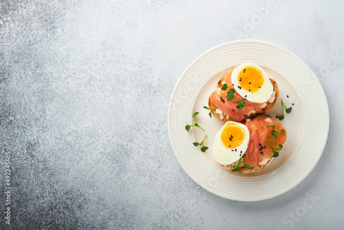 Sandwich with delicious toppings on top: smoked salmon, eggs, herbs and microgreens radish, black sesame seeds over white plate on concrete background. Healthy open sandwich superfood. Top view.
