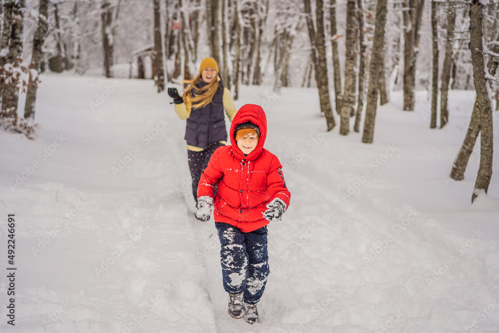 Funny little boy and his mom in blue winter clothes walks during a snowfall. Outdoors winter activities for kids. Cute child wearing a warm hat low over his eyes catching snowflakes with his tongue