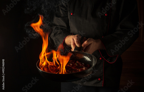 Chef prepares vegetable food in a frying pan with a flame of fire. The concept of restaurant and hotel service. Flamber or cooking over natural fire. Grande cuisine photo