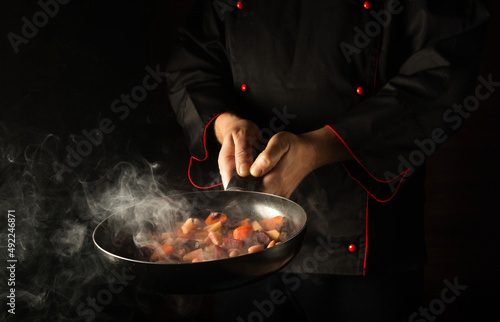 Fototapeta Naklejka Na Ścianę i Meble -  Professional chef prepares food in a frying pan with steam on a black background. The concept of restaurant and hotel service. European cuisine