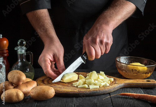 Cook cuts raw potatoes into pieces with a knife before preparing breakfast or dinner. Close-up of chef hands while working in kitchen