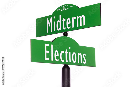 Closeup of midterm elections crossroads green street sign with white text atop black road sign pole showing intersection of 2022 midterm and elections against white background. photo