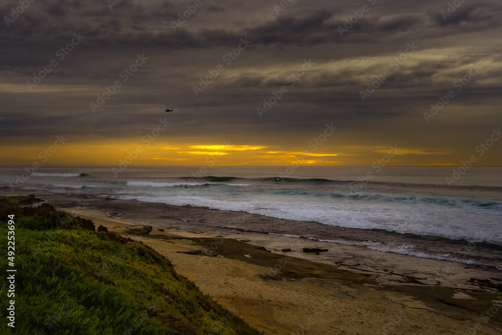 2022-03-12 THE LAJOLLA SHORELINE WITH A SAND AND ROCK SHORELINE AND NICE WAVES, SURFERS AND A HELICOPTER IN THE DARK SKY WITH A BEAUTIFUL SUNSET PEEKING THROUGH. 2022-03-12 THE LAJOLLA SHORELINE WITH A SAND AND ROCK SHORELINE AND NICE WAVES, SURFERS AND A HELICOPTER IN THE DARK SKY WITH A BEAUTIFUL SUNSET PEEKING THROUGH.