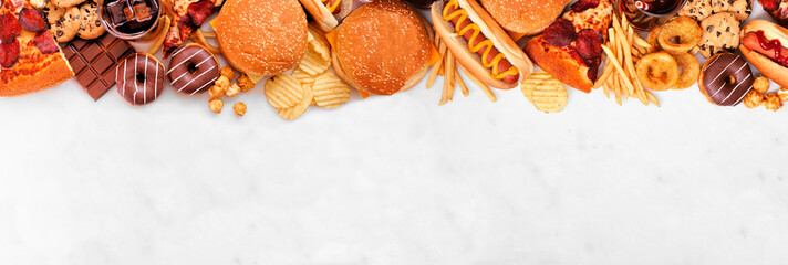Junk food top border over a white marble banner background. Selection of take out and fast foods. Pizza, hamburgers, french fries, chips, hot dogs, sweets. Top down view with copy space.
