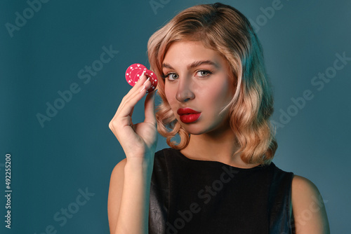 Blonde girl in black leather dress showing two red chips  posing against blue background. Gambling entertainment  poker  casino. Close-up.