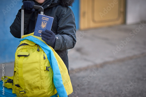 Evacuation of civilians, sad child with the flag of Ukraine. Refugee family from Ukraine crossing the border. Hand holding a passport above the luggage with yellow-blue flag. Stop war, support Ukraine