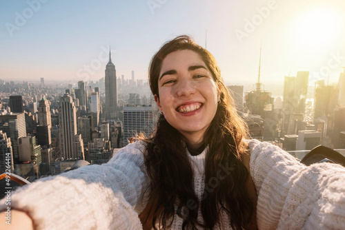 Young woman taking a selfie smiling on a rooftop with the empire state building in the background during sunset. Travel concept. influencer concept. Happiness concept. photo