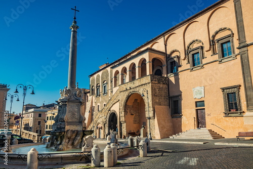 Tarquinia, Viterbo, Lazio, Italy - The main square of the village. The circular fountain with the obelisk and the cross. The town hall illuminated by the sun, in the blue sky.