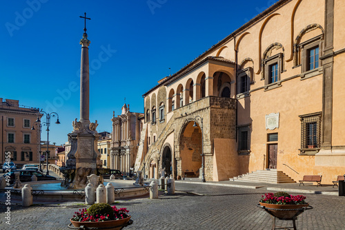Tarquinia, Viterbo, Lazio, Italy - The main square of the village. The circular fountain with the obelisk and the cross. The town hall illuminated by the sun, in the blue sky.
