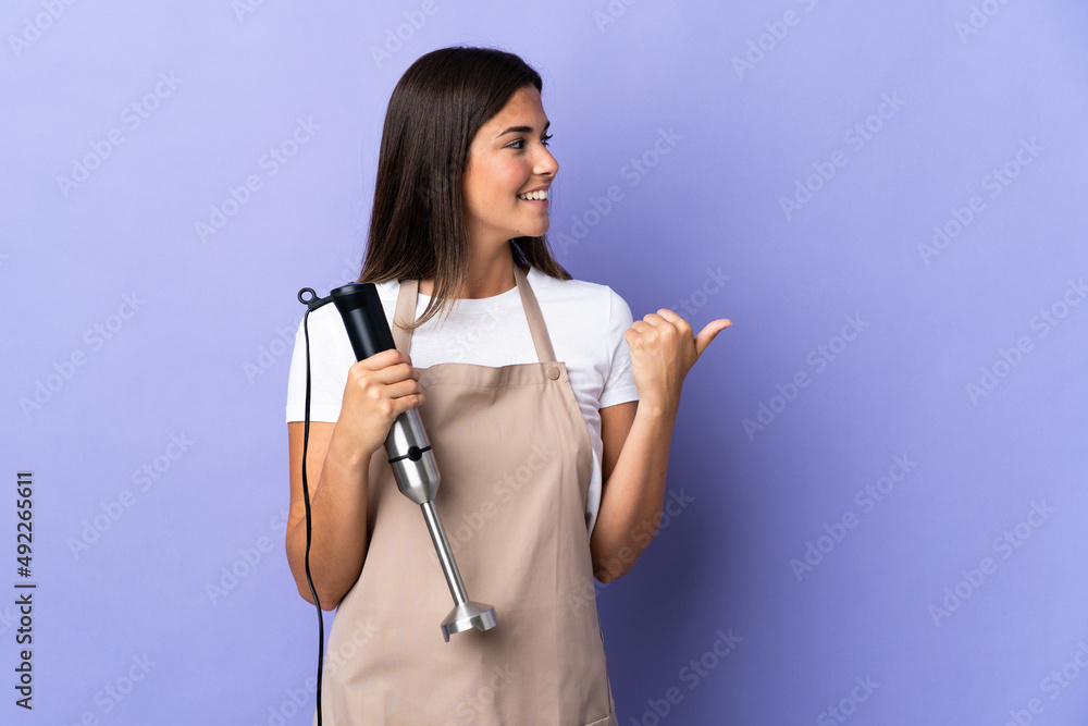 Brazilian woman using hand blender isolated on purple background pointing to the side to present a product Brazilian woman using hand blender isolated on purple background pointing to the side to present a product