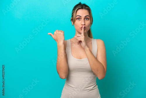 Young caucasian woman isolated on blue background pointing to the side and doing silence gesture