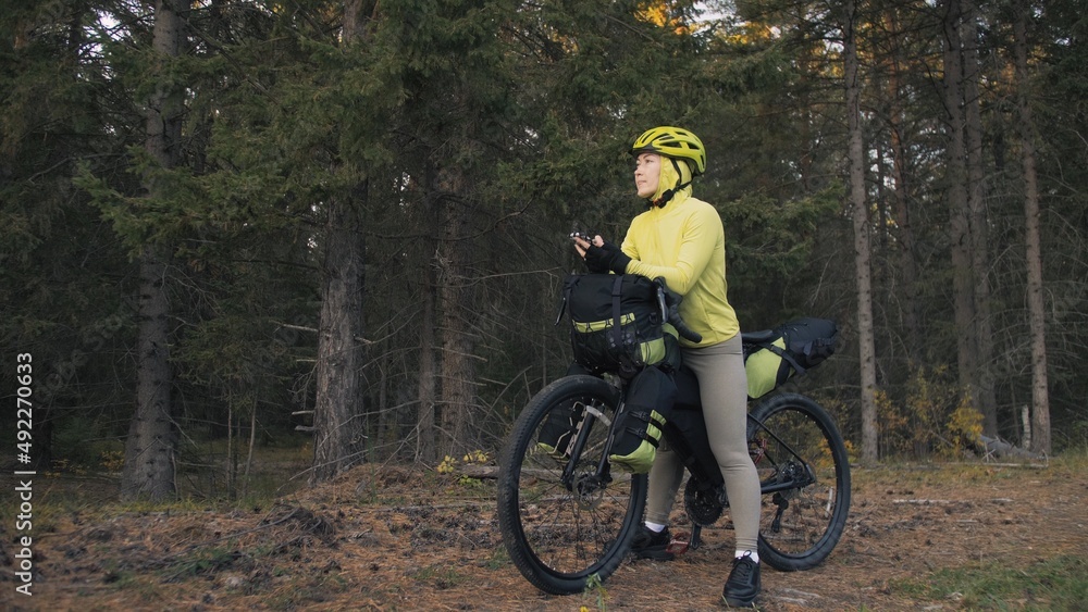 The woman travel on mixed terrain cycle touring with bike bikepacking outdoor. The traveler journey with bicycle bags. Sportswear in green black colors. Magic forest park. Make a selfie smartphone.