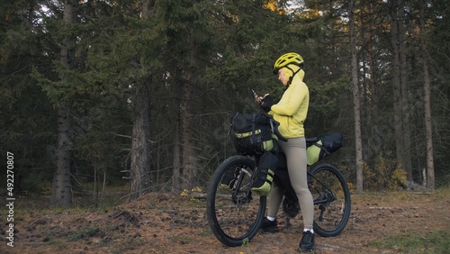 The woman travel on mixed terrain cycle touring with bike bikepacking outdoor. The traveler journey with bicycle bags. Sportswear in green black colors. Magic forest park. Make a selfie smartphone.