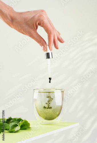 Female hand pours liquid chlorophyll into a glass of water with a dropper. Liquid chlorophyl and fresh herbs on the green table. Green background, natural sunlight, vertical. Superfood and healthy