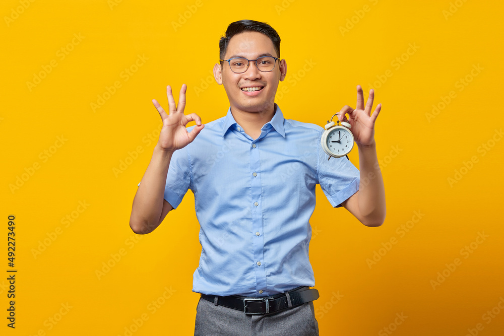 Portrait of cheerful handsome Asian young man in glasses showing alarm clock and showing okay gesture with finger isolated on yellow background. Time management lifestyle concept Portrait of cheerful handsome Asian young man in glasses showing alarm clock and showing okay gesture with finger isolated on yellow background. Time management lifestyle concept
