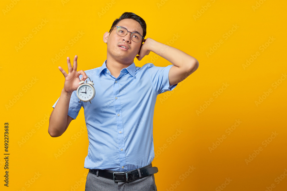 Pensive handsome Asian young man in glasses showing alarm clock and looking up isolated on yellow background. Time management lifestyle concept Pensive handsome Asian young man in glasses showing alarm clock and looking up isolated on yellow background. Time management lifestyle concept