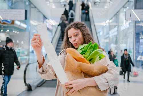 Rising food prices. Surprised woman looking into a paper check at the mall, holding a paper bag with fresh herbs and a baguette.