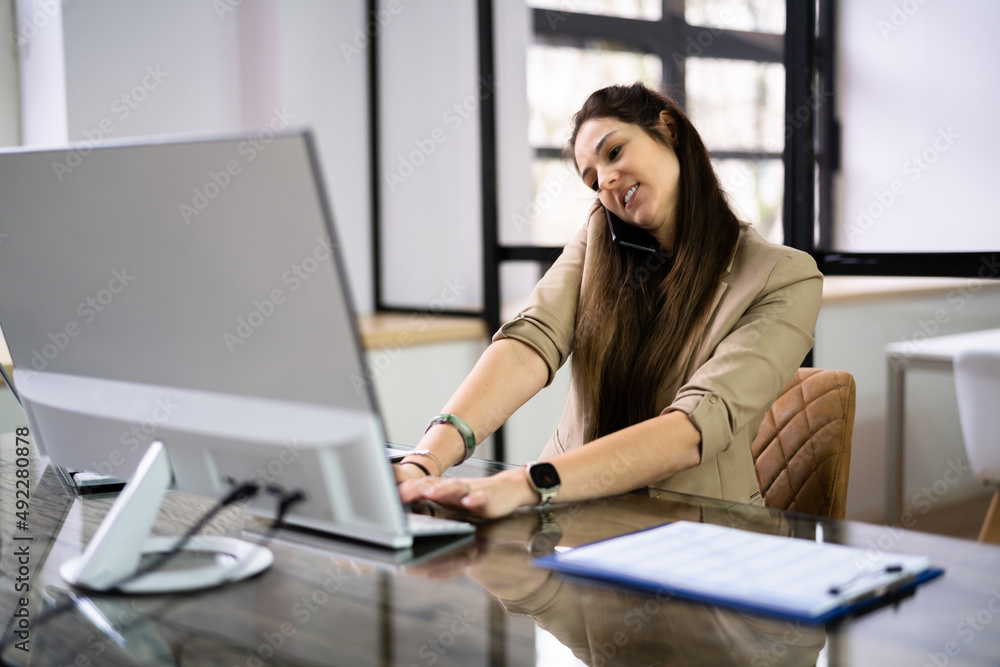 Young Businesswoman On Call While Using Computer At Desk Young Businesswoman On Call While Using Computer At Desk