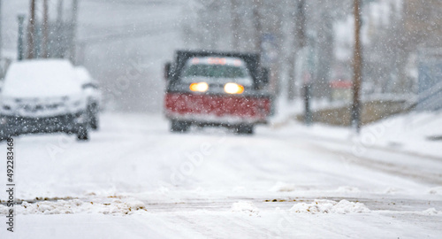 Snow storm plow, small USA town road covered in snow, during snow storm, cars traffic, bad weather travel.