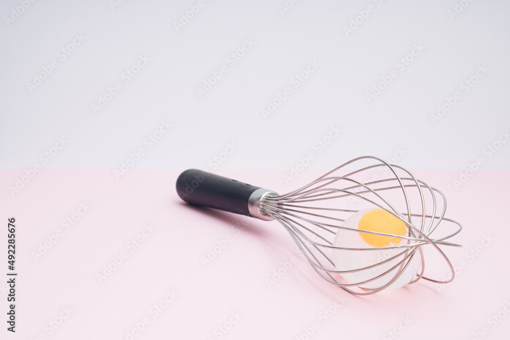Whisk with transparent egg on pastel pink. Light banner background, copy space for text. Culinary mixer, whipping egg white and yolk, preparing and mixing cream. Whisk with transparent egg on pastel pink. Light banner background, copy space for text. Culinary mixer, whipping egg white and yolk, preparing and mixing cream.