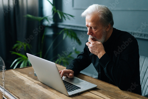 Close-up of thoughtful worried senior aged businessman working on laptop at workplace in dark home office room. Side view of concentrated mature adult male using computer looking on screen. photo
