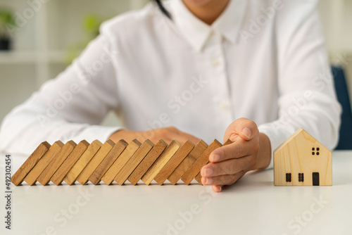 A woman's hand stops a wooden block from falling onto a model house. Home insurance and safety ideas and protecting your home from falling over wooden blocks.