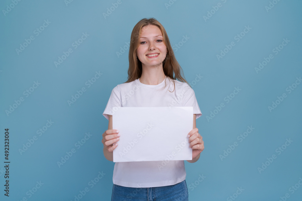 Young woman holding white paper on blue background. Caucasian woman with white blank template sheet with empty space in white t-shirt and blue jeans shows advertisement, mockup Young woman holding white paper on blue background. Caucasian woman with white blank template sheet with empty space in white t-shirt and blue jeans shows advertisement, mockup