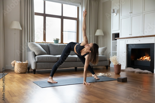 Young attractive woman wearing sportswear doing Utthita Trikonasana pose Extended Triangle asana working out alone in living room. Yoga practice at modern cozy home, active healthy lifestyle concept