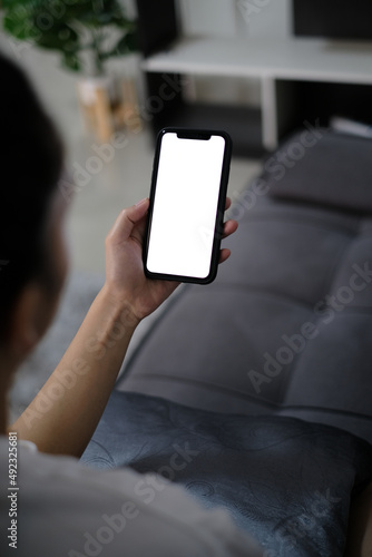 asia women holding smart phone with mockup white blank display, empty screen for app ads sitting on couch at home. Mobile applications technology concept, over shoulder close up view.