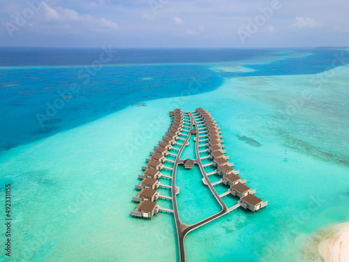 Overwater villas and white sand beach on tropical island for holidays vacation travel and honeymoon. Luxury resort hotel in Maldives or Caribbean with turquoise sea water. Drone aerial view.