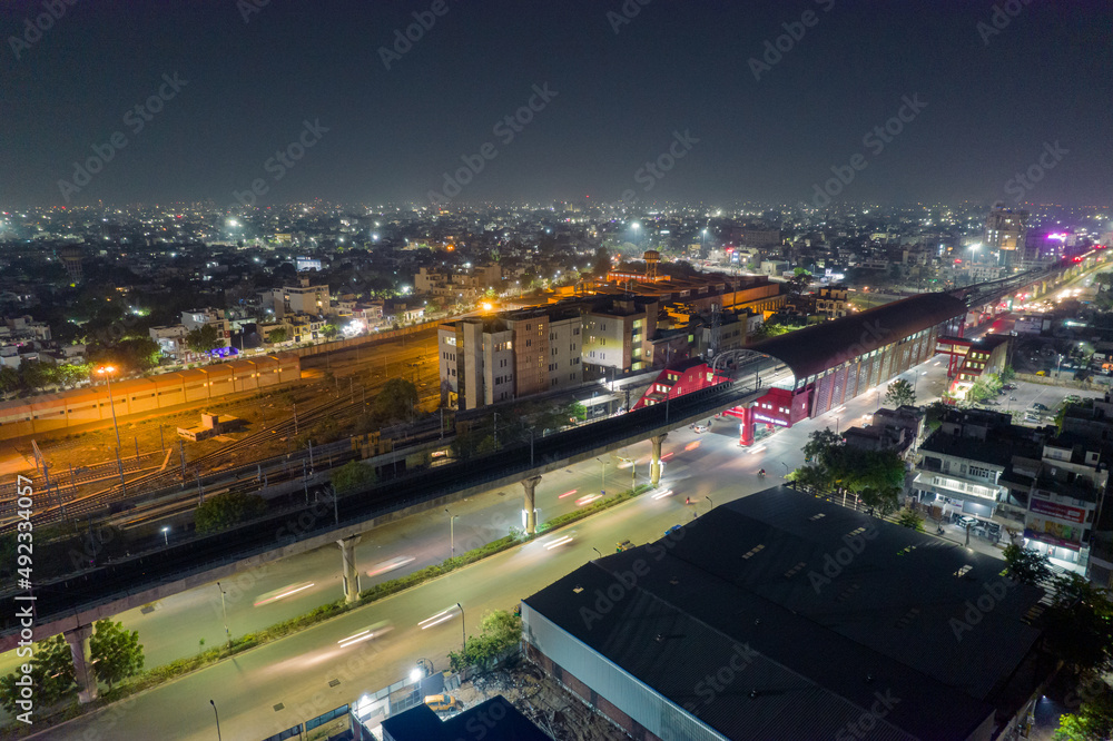 elevated metro track station with train leaving and light trails over lit busy street and yard in distance showing rapid development of Indian cities